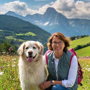 Eine Frau und ein weißer Hund sitzen auf einer Wiese mit Bergkulisse im Hintergrund.
