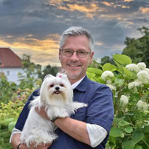 Ein lächelnder Mann hält einen weißen, flauschigen Hund mit einer rosa Schleife vor einem Hintergrund aus bewölktem Himmel und viel Grün.